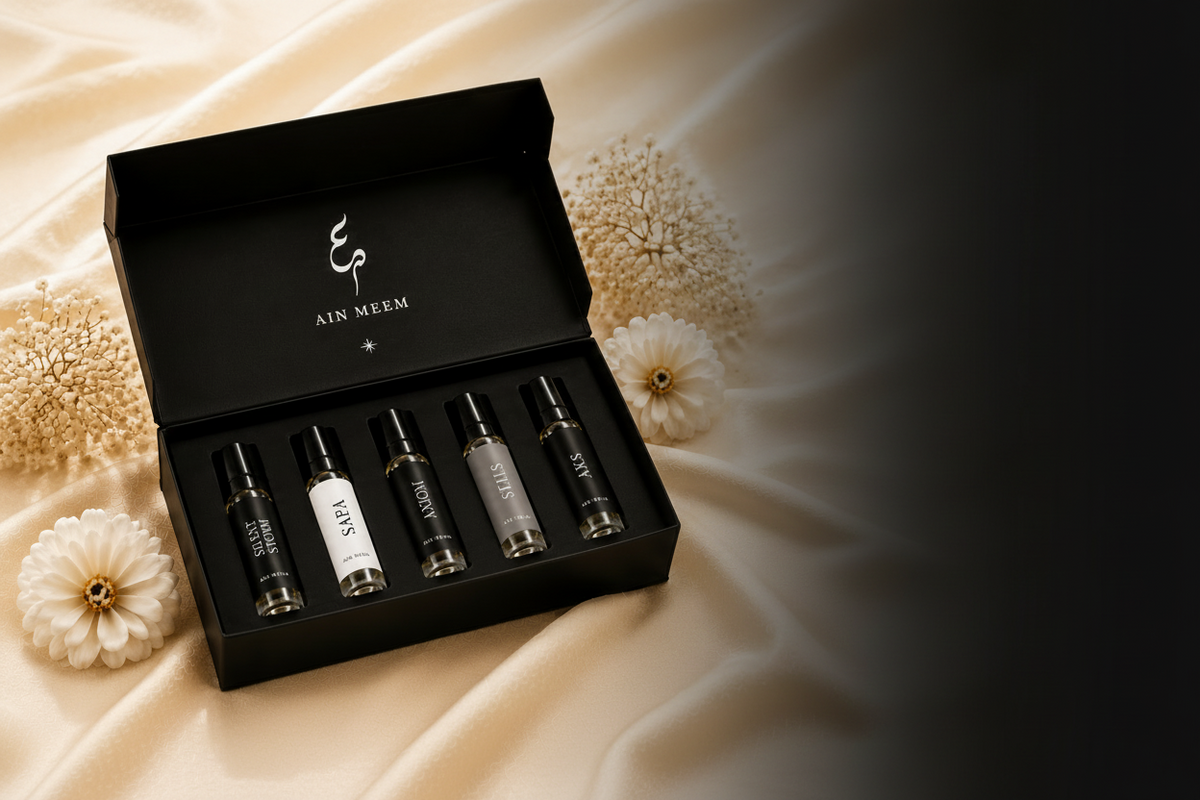A black gift box containing five small cosmetic sample vials, displayed on a cream satin fabric with decorative dried flowers. The brand name 'ALI MEEM' is visible on the inside lid.